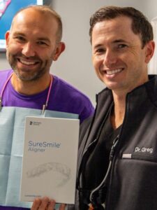 Two smiling dentists, one in a purple shirt and the other in a black jacket labeled Dr. Gregory Shvartsman, hold a SureSmile Aligner box in a dental office.