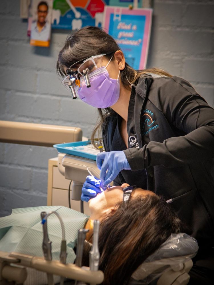 Dr. Gloria Luong, wearing a face shield, mask, and gloves, examines a patient’s mouth in a dental clinic. Dental tools and informational brochures are visible in the background.