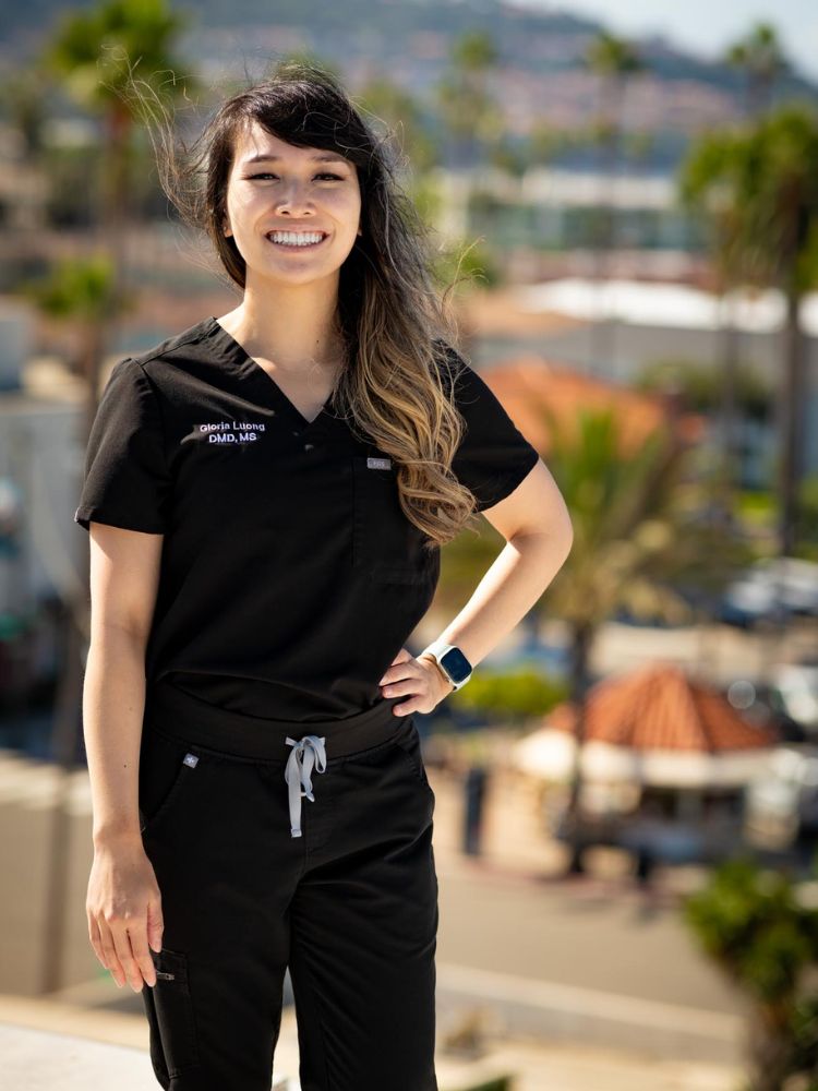 Dr. Gloria Luong, smiling in black medical scrubs, stands outdoors with one hand on her hip. Palm trees, buildings, and a sunny, blurred background suggest a warm, urban location.