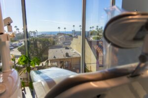 A dental chair at South Redondo Dental sits in a bright office, facing large windows with views of buildings, palm trees, and the ocean under a clear blue sky.