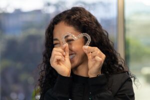 A woman with curly hair smiles while holding a clear dental aligner in the shape of a heart in front of her face. She stands indoors with a blurred background, highlighting her happiness with South Redondo Dental care.