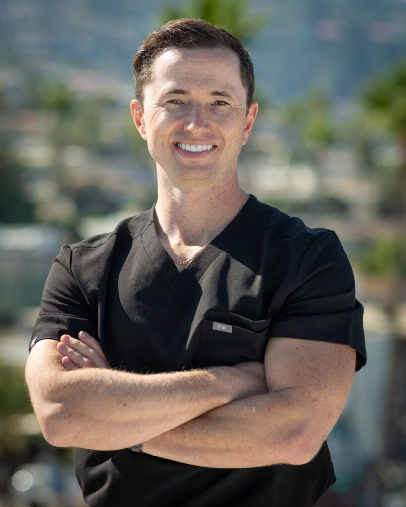 Dr. Gregory Shvartsman, a smiling man with short brown hair, stands outdoors with his arms crossed, wearing black medical scrubs. The background is blurred, showing greenery and buildings.