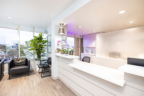 Modern office reception area at South Redondo Dental with a white desk, potted plants, black chairs, and large windows letting in natural light; outside, trees and parked cars are visible.