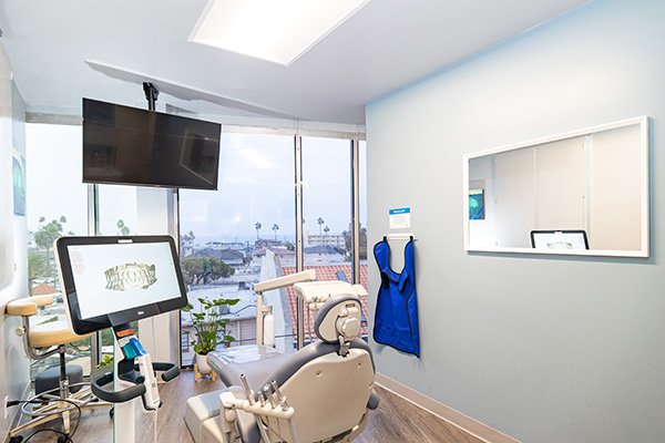A bright dental exam room at South Redondo Dental features a modern chair, monitor with dental scans, wall-mounted TV, lead apron, and large windows showcasing city views. Light blue walls and a mirror add to the welcoming atmosphere.