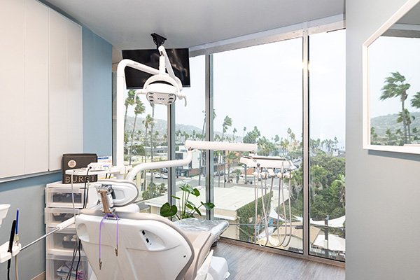 A modern dental office at South Redondo Dental features a dental chair facing floor-to-ceiling windows overlooking a cityscape with palm trees and hills. The bright, clean room has blue walls and sleek white cabinets.