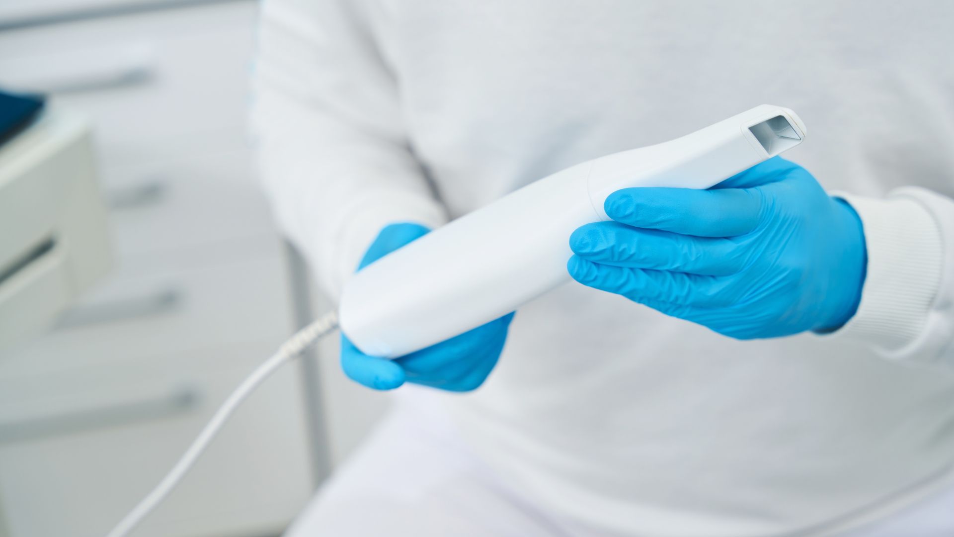 A person wearing blue gloves holds a white intra-oral camera or similar handheld medical device, with a blurred clinical background.