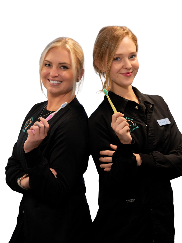 Two women in black uniforms stand back-to-back, each smiling and holding a toothbrush. They appear confident and professional, suggesting expertise in teeth cleaning or dental hygiene. The background is plain white.