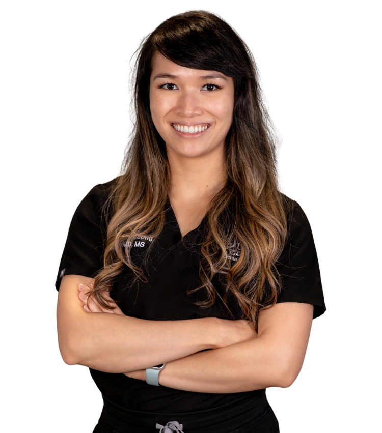 A woman with long, wavy brown hair stands smiling with arms crossed, wearing black medical scrubs and a smartwatch, representing Home Care services against a plain white background.