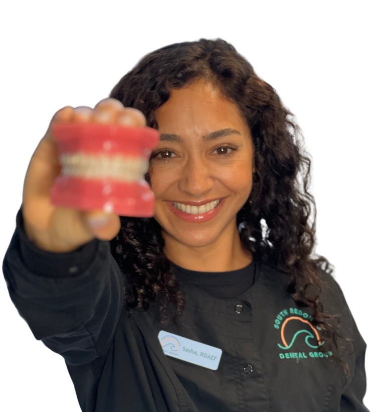 A smiling woman with curly hair in a dental uniform holds a model of teeth toward the camera, representing Periodontal Therapy, standing against a plain white background.