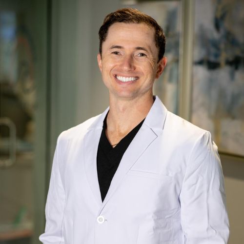 A smiling Dr. Gregory Shvartsman in white lab coats stand blur office in the background. Confident and professional vibe.