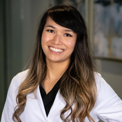 A woman with long, wavy dark hair and blonde highlights smiles at the camera. Wearing a white lab coat, she stands indoors at South Redondo Dental, with blurred artwork visible in the background.