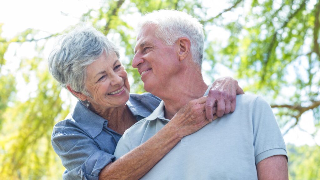 An elderly couple with missing teeth smiles lovingly at each other outdoors. The woman hugs the man from behind, and both appear happy and content in the sunlight, with green trees in the background.