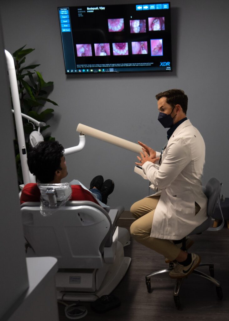 A dentist wearing a mask talks to a patient in a dental chair while pointing to dental X-ray images displayed on a monitor on the wall.