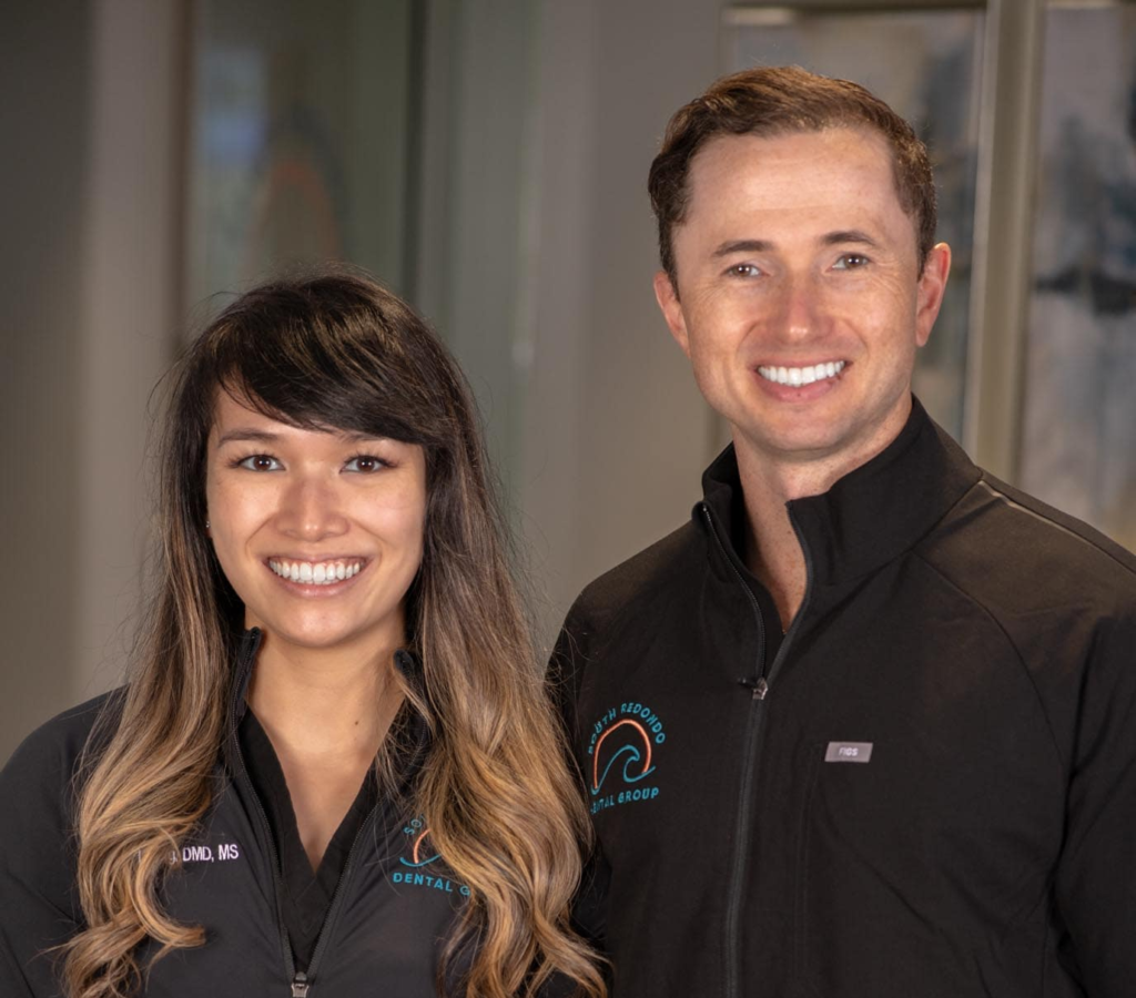Two people wearing black zip-up jackets with embroidered dental group logos smile while standing indoors. The woman has long, wavy hair; the man has short, brown hair. Both appear friendly and professional.