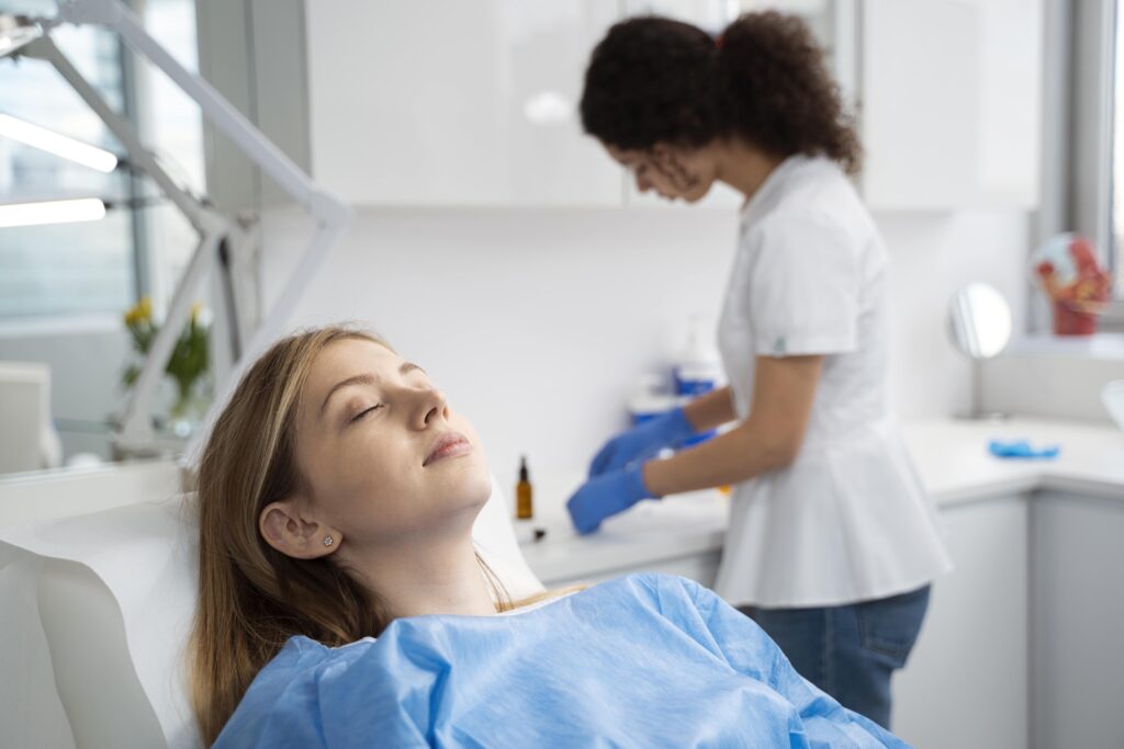 A woman in a medical gown lies on a reclining chair with her eyes closed, while a healthcare professional in gloves prepares tools at a counter in a bright, modern clinic.