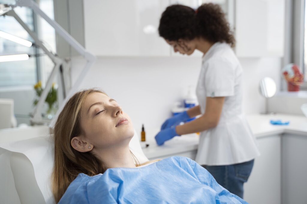 A woman in a blue medical gown relaxes with her eyes closed on a chair, while a healthcare professional wearing gloves prepares supplies at a counter in a bright clinic room.