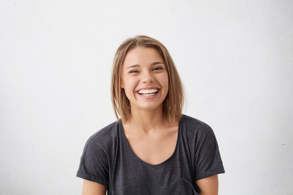 A young woman with straight, shoulder-length light brown hair smiles broadly, showing her teeth. She is wearing a dark gray T-shirt and is posed in front of a plain white background.